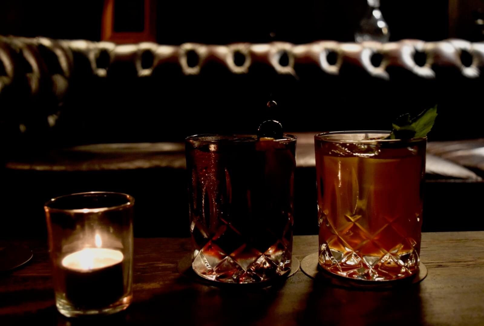 Two whisky cocktails on a table in front of a leather couch 