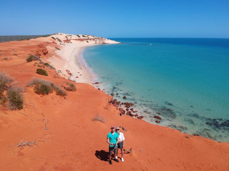 Hayley and Enrico standing on red sand with a white beach and turquoise water in the background 