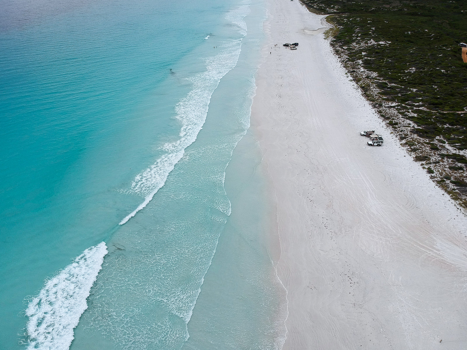 A drone shot of 4WD vehicles parked on a white sand beach with turquoise ocean