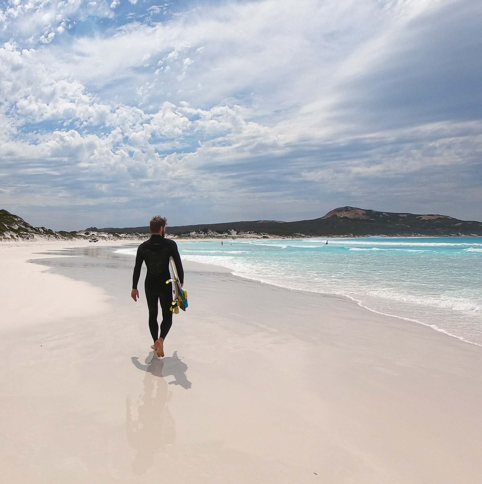 A surfer walking along a white sand beach with turquoise waves lapping the shore 