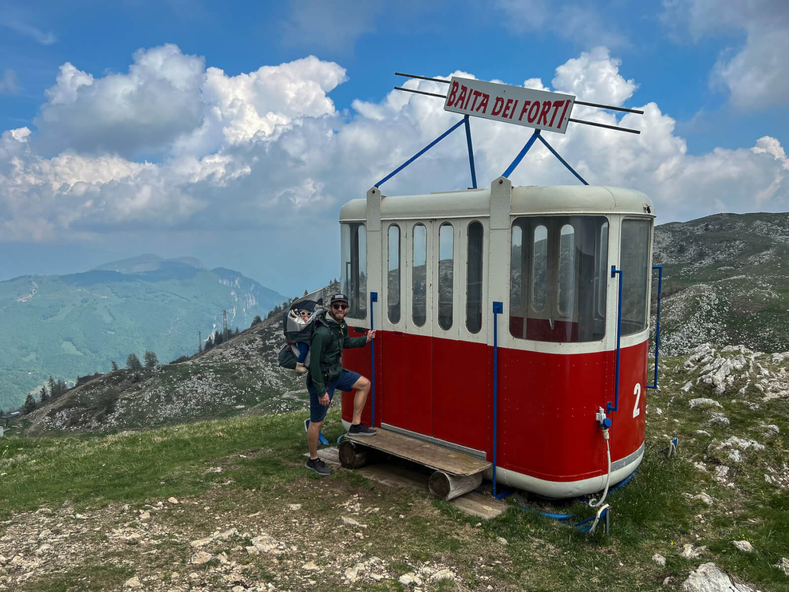 A man and baby hiking on  Monte Baldo, standing next to an old cable car.