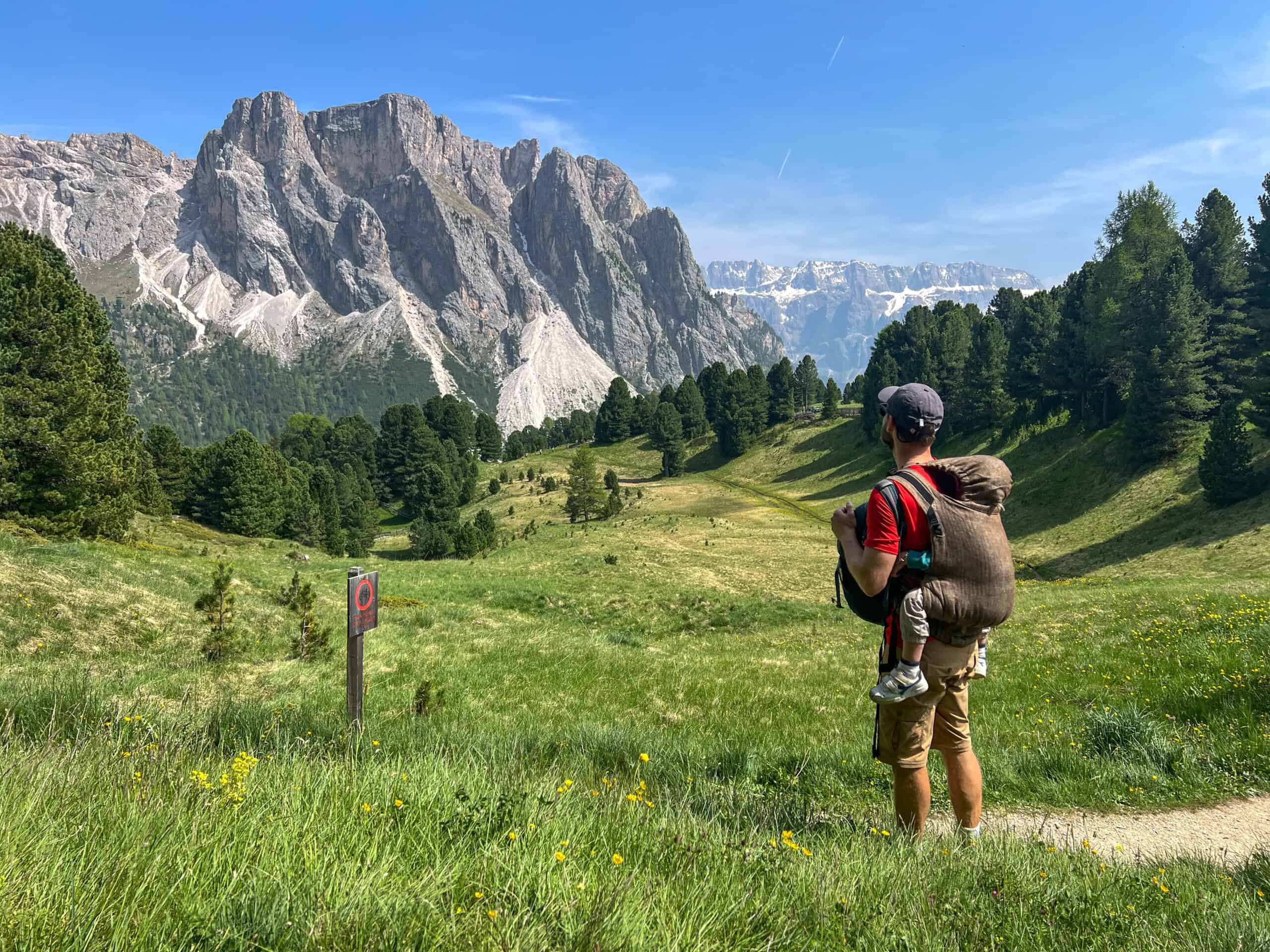 A father carries his young son in a baby carrier while hiking along a scenic mountain trail in the Dolomites.