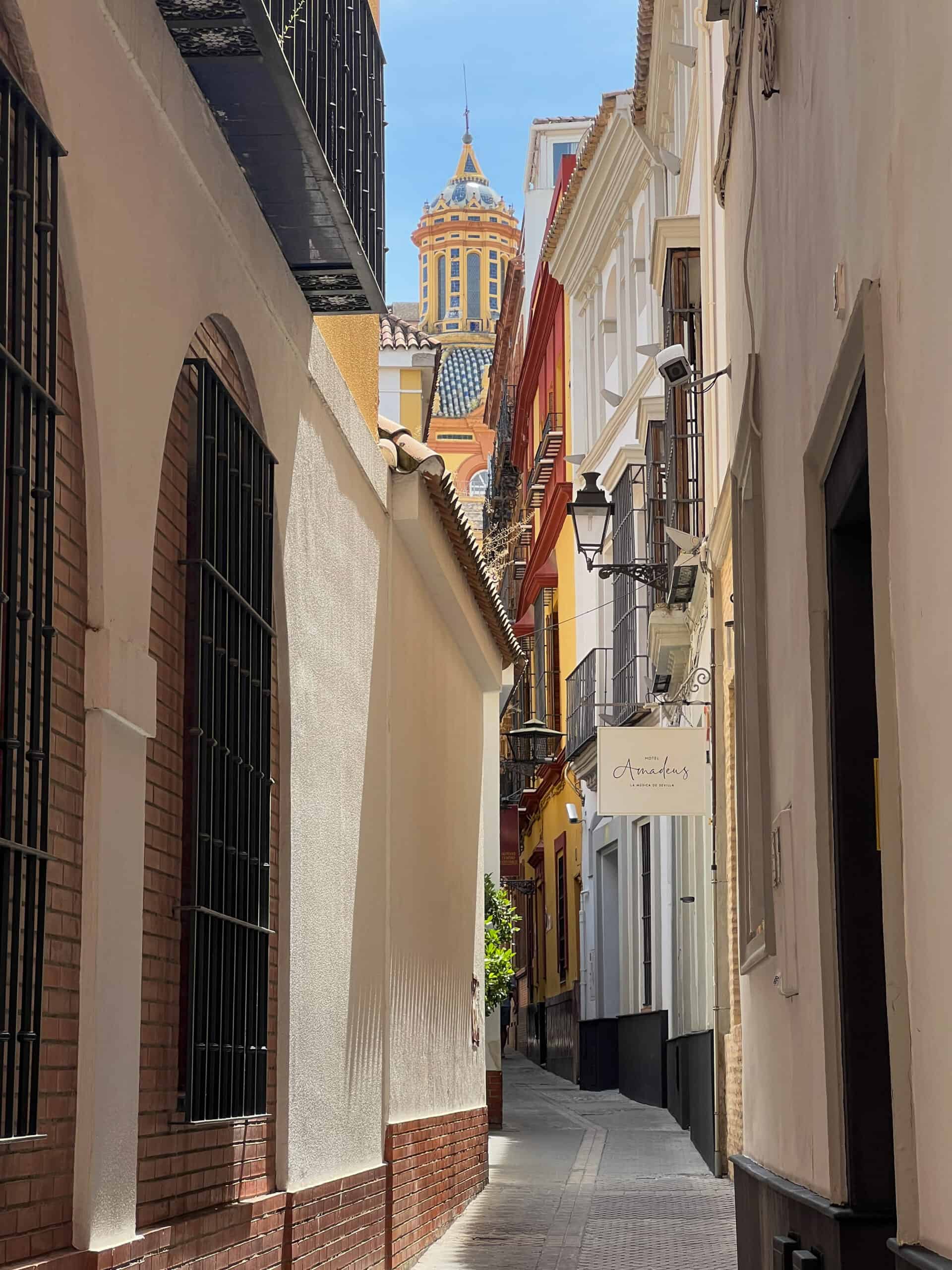 Sunlit alley in Seville framed by white-washed walls and a colourful tiled dome in the distance.
