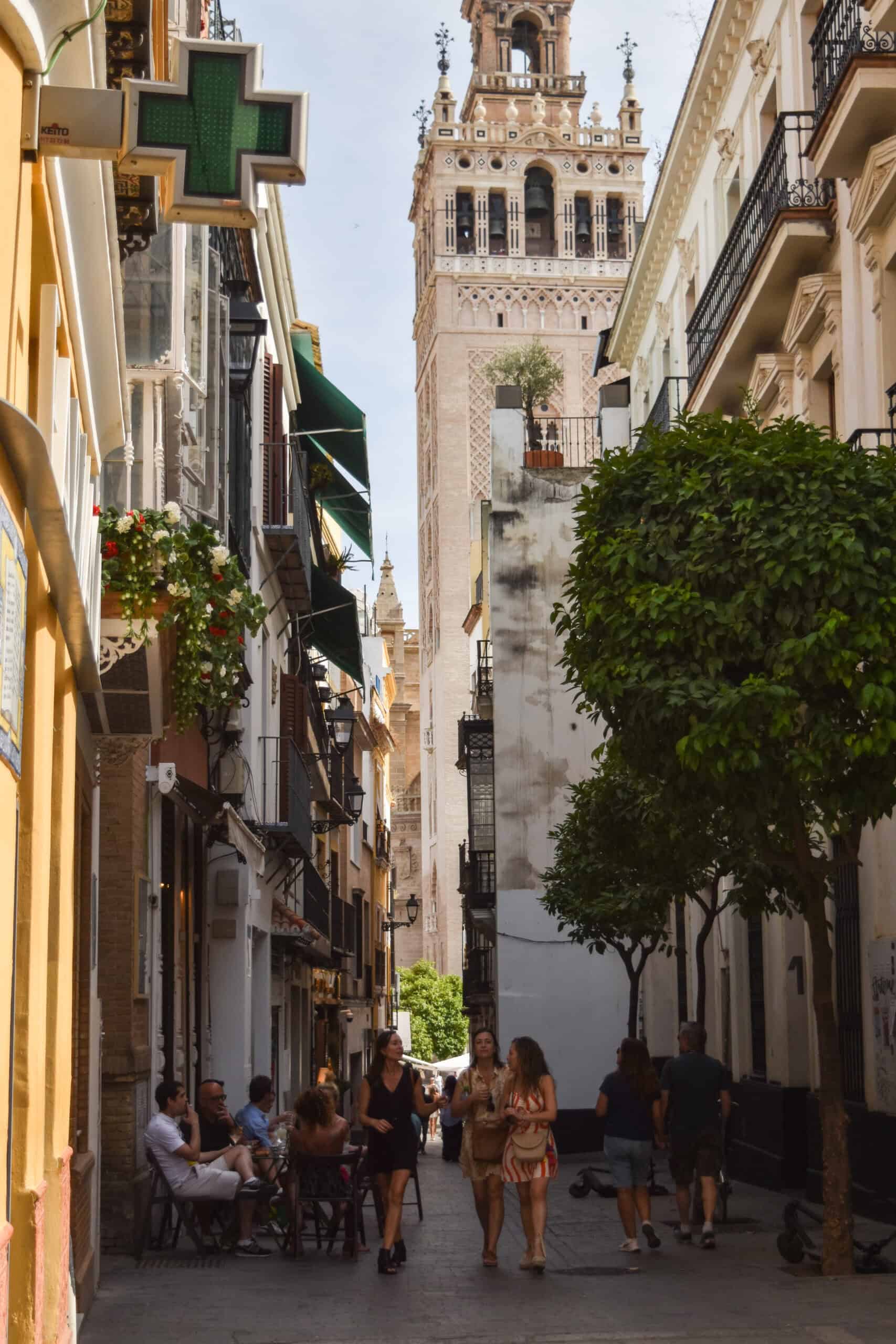 Narrow Seville street with the Giralda tower rising above cafés and walking locals.
