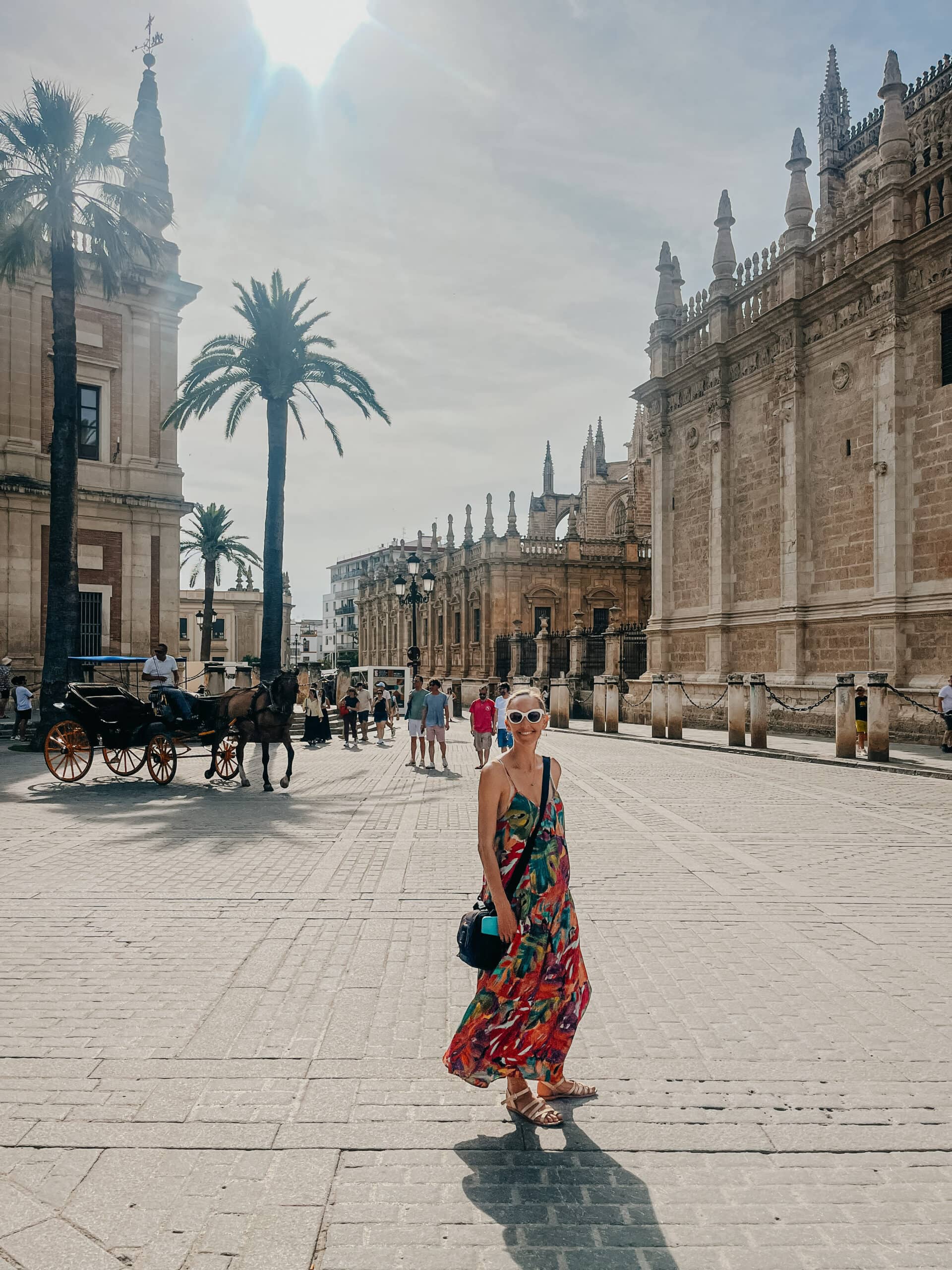 A woman standing in the plaza beside Seville Cathedral with a horse-drawn carriage in view.