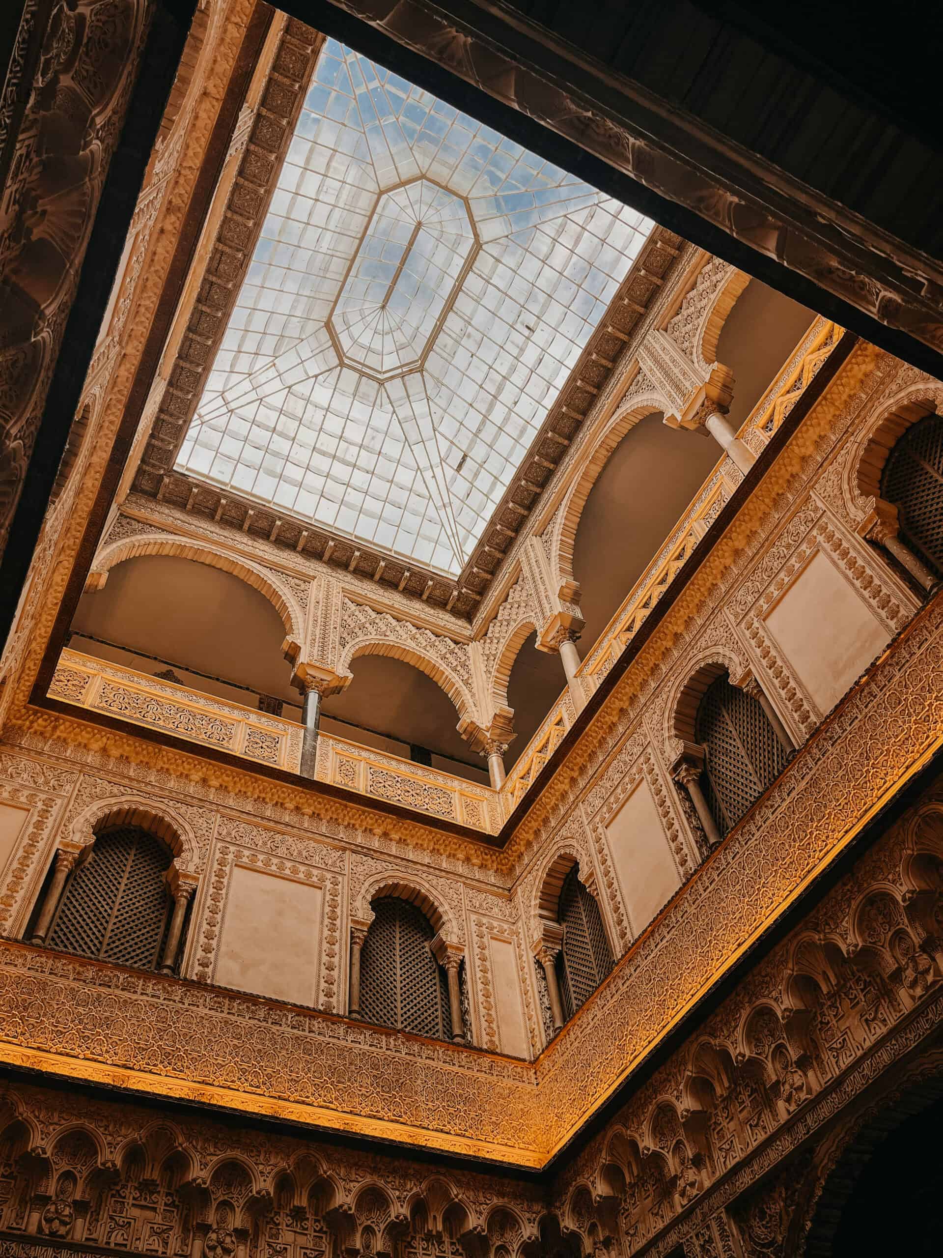 Alcázar interior looking up at an ornate glass skylight and carved plaster arches.