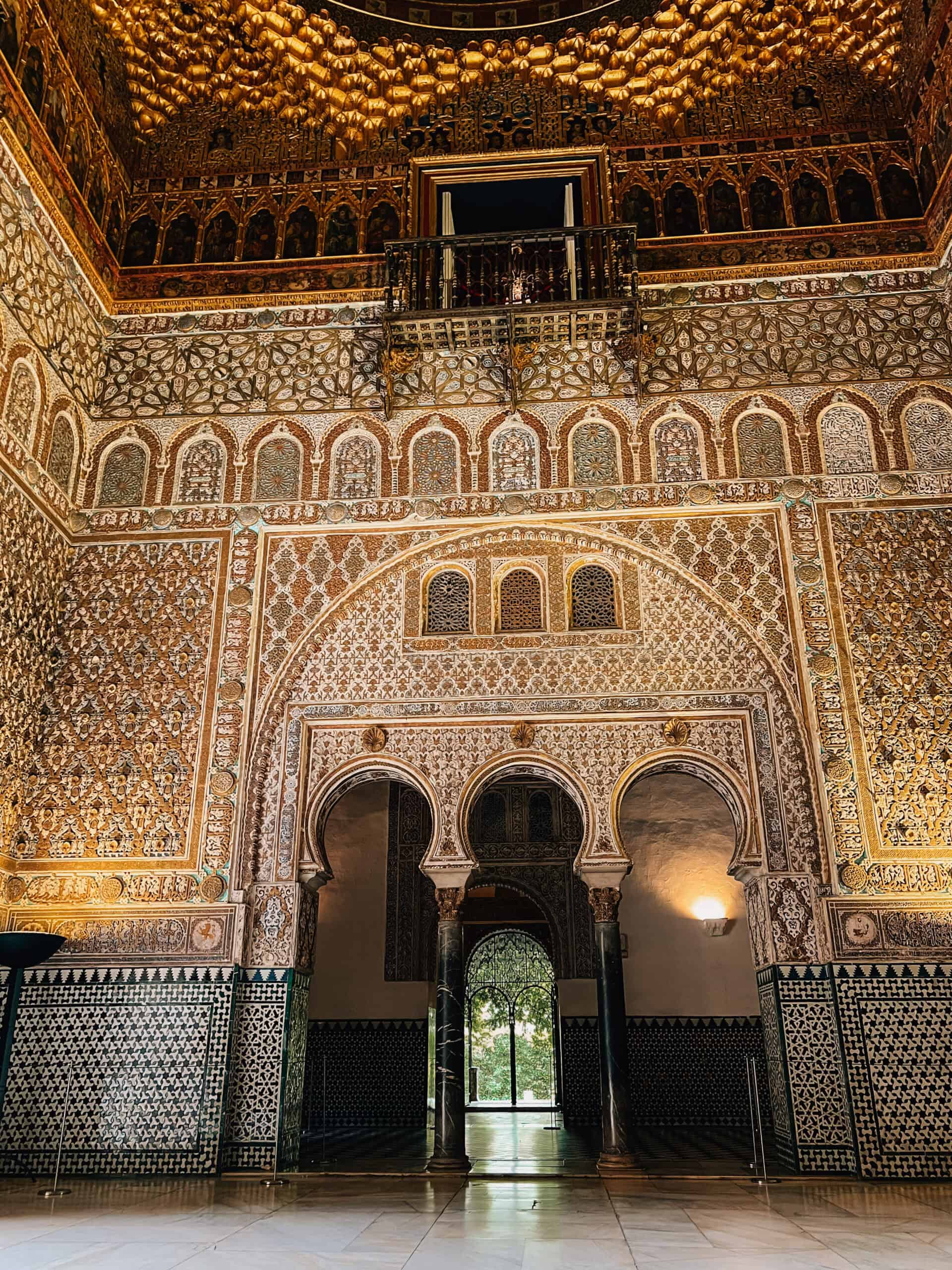Golden-lit ornate hall inside the Alcázar with intricate carved detail and arched doorway.