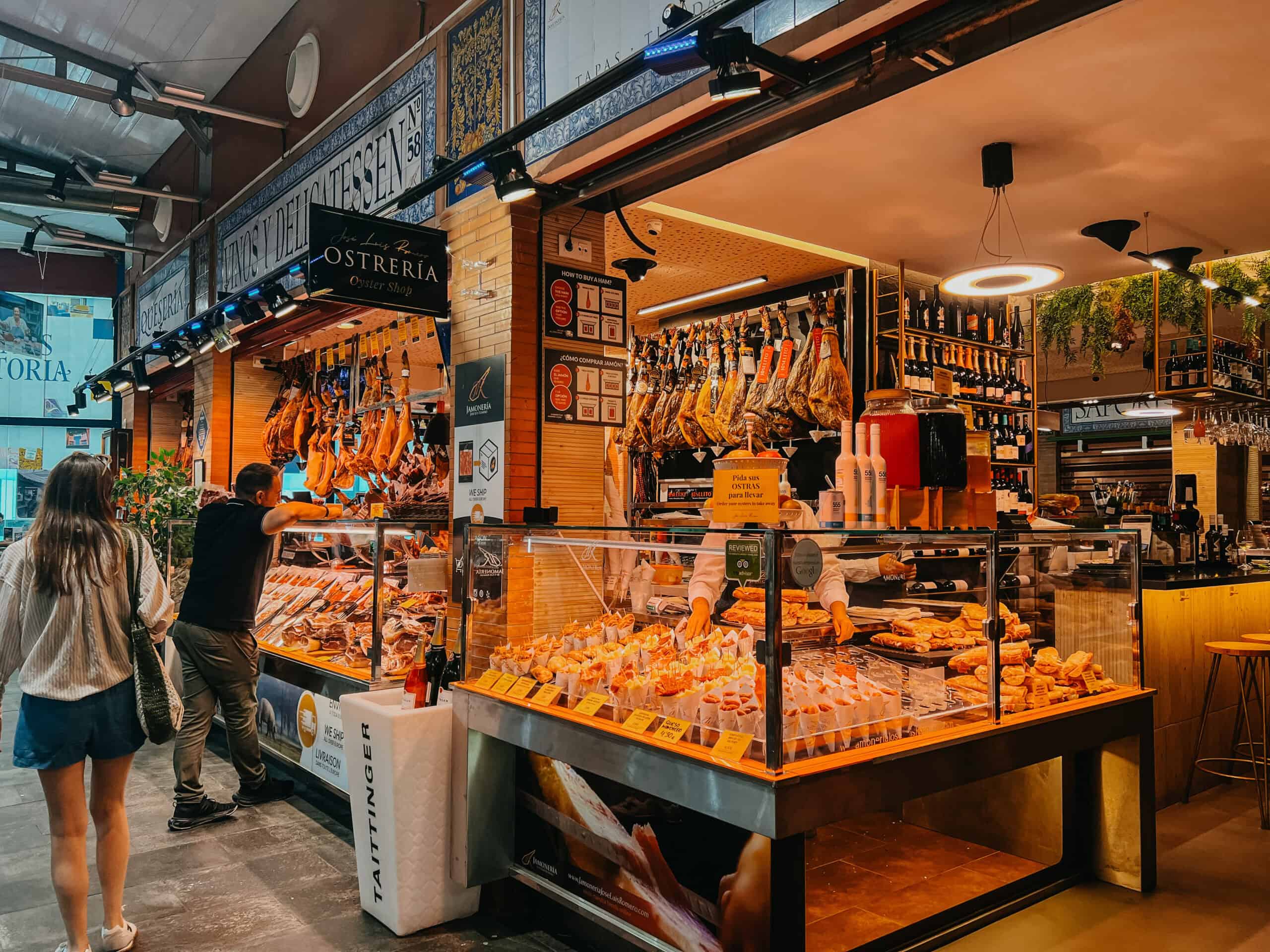 Busy market stall displaying hanging jamón, tapas cups and fresh produce inside Mercado de Triana.